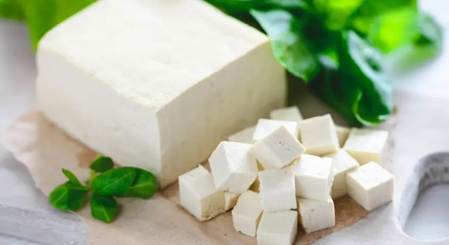 Kostka i pokrojone w kostkę tofu, obok zielone listki. Tak wygląda tofu – białe, delikatne, gotowe do przygotowania.