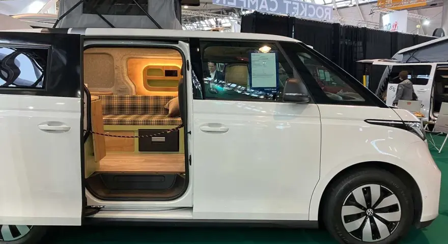 Vit ID Buzz camper van med uppfällt tak och öppen skjutdörr som visar en mysig interiör med rutig soffa och trädetaljer.