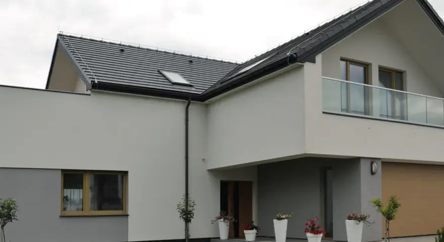 Nowoczesny dom z kremową elewacją, szarym cokołem, dużym oknem i balkonem ze szklaną balustradą. Przed wejściem białe donice z kwiatami.