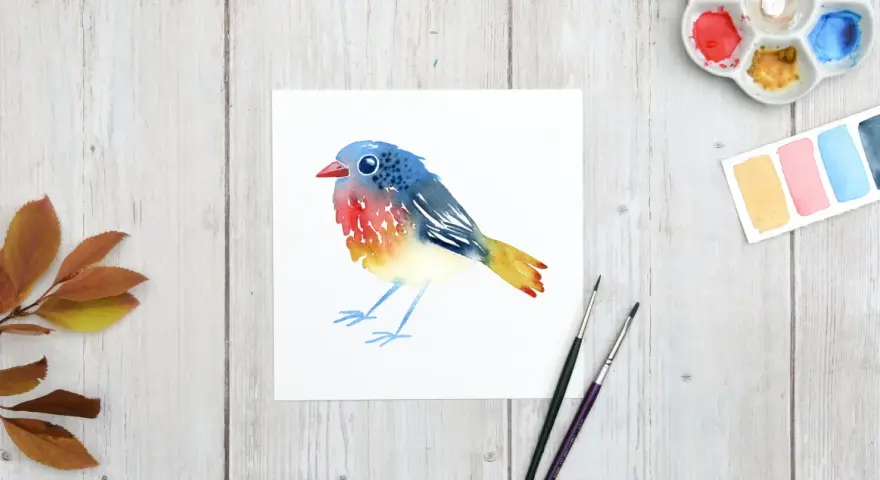 Un charmant dessin oiseau aquarelle aux couleurs vives, posé sur du papier blanc. Des feuilles d'automne et du matériel de peinture complètent la scène.