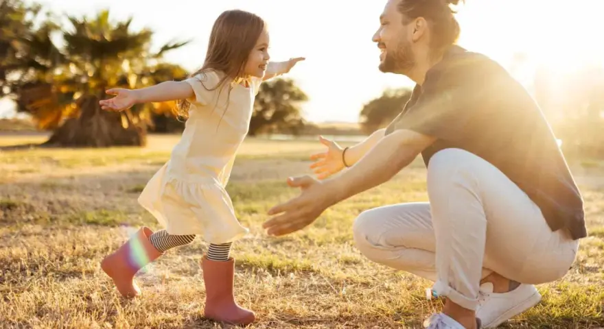 Ein kleines Mädchen rennt mit offenen Armen auf ihren lachenden Vater zu. Die warme Abendsonne beleuchtet die Szene und unterstreicht die liebevolle vater tochter beziehung.