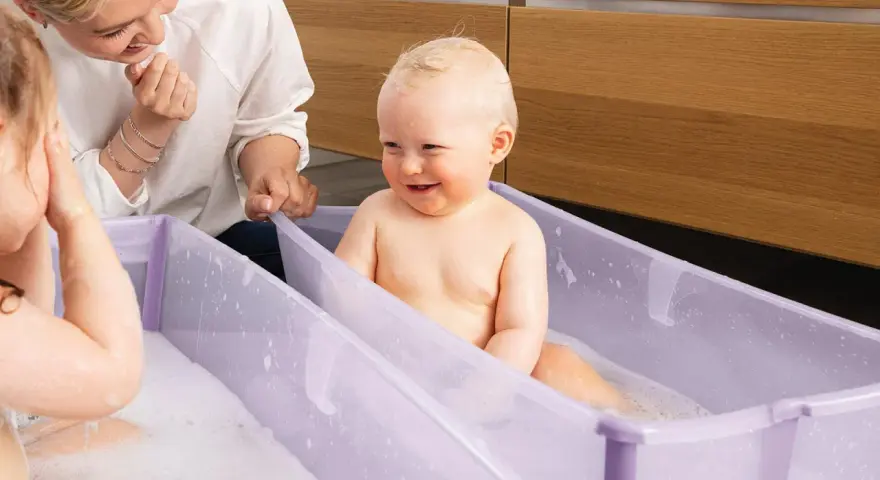 Uśmiechnięty bobas w fioletowym krzesełku do kąpieli, mama patrzy z czułością.