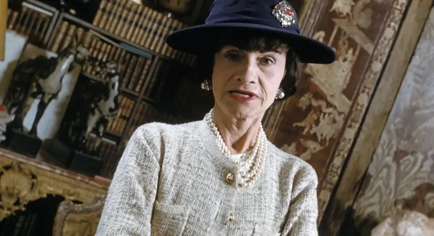 Iconica stilista Coco Chanel, con cappello blu e perle, in un ambiente elegante con libri e decorazioni.