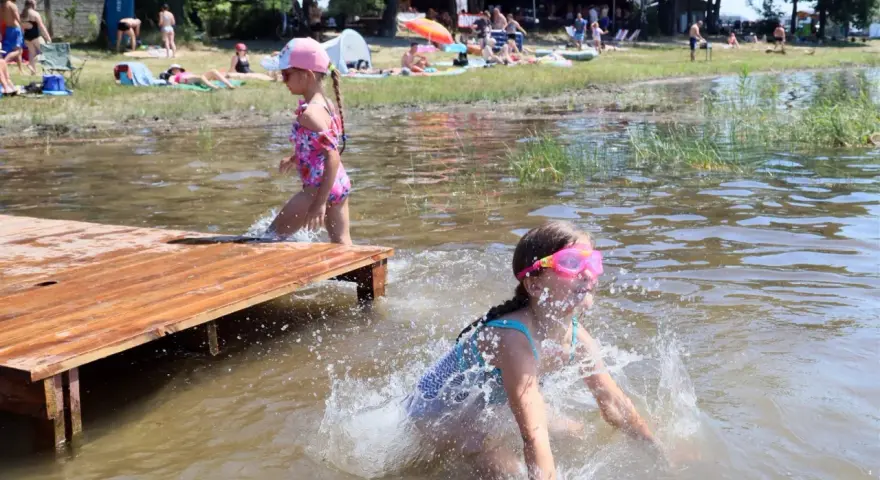 Dwie dziewczynki pluskają się w wodzie przy drewnianym pomoście. W tle plażowicze i drzewa. Wakacje nad jeziora lubelskie.