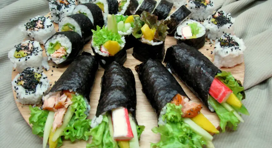 Talerz sushi z futomaki i uramaki. Widać rolki z łososiem, awokado, sałatą, paluszkami krabowymi i mango.
