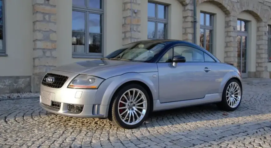 Silberner Audi TT 2005 mit schwarzem Dach parkt auf Kopfsteinpflaster vor einem Gebäude mit Steinbögen.