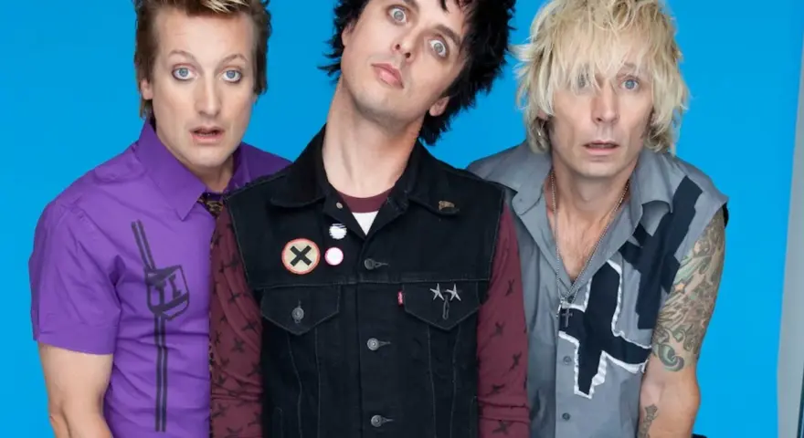 Trzech członków zespołu Green Day Polska z wyrazistymi minami na niebieskim tle.
