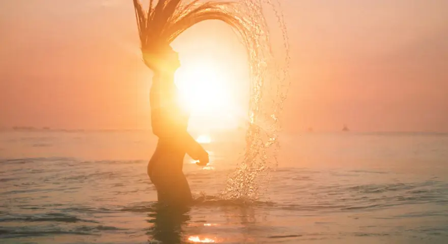 Silhouette d'une femme jetant ses cheveux dans l'eau au coucher du soleil, avec un effet de lens flare photoshop.
