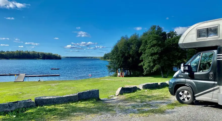 Husbil vid en sjö med brygga, perfekt för fricamping Sverige. Blå himmel och grönska.