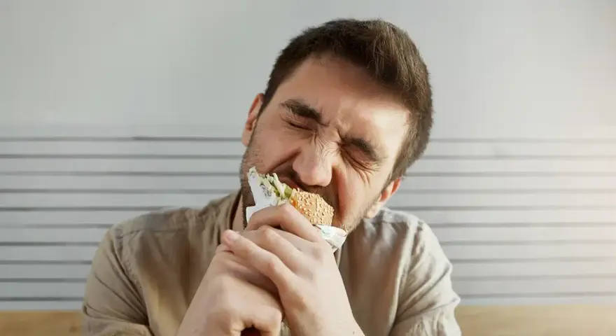 Uomo con espressione sofferente mangia un panino, forse per alleviare lo stress, ma teme di ingrassare.