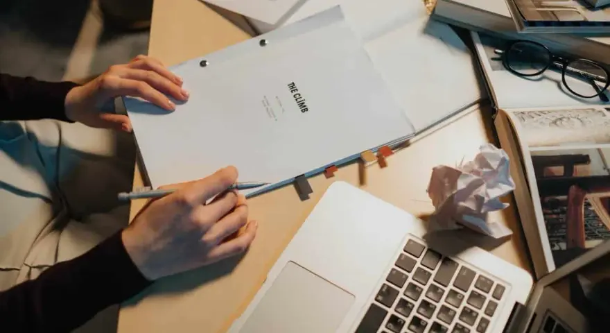 Manos escribiendo un guion cortometraje, rodeado de libros, gafas y un portátil.