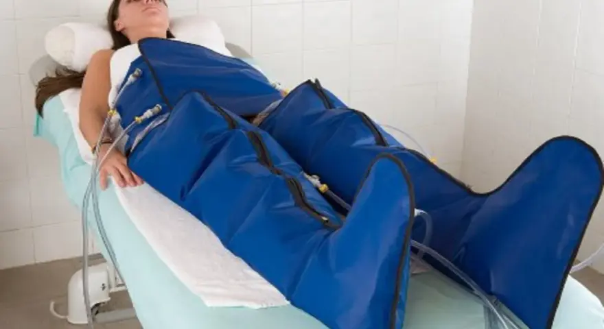 Kobieta leży na łóżku, jej nogi są owinięte w niebieskie mankiety urządzenia do mechanicznego drenażu limfatycznego.
