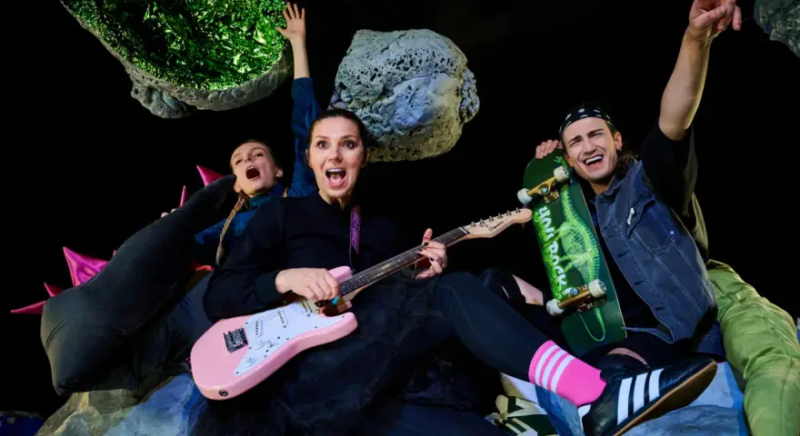 Energiczna ekipa w kosmicznej scenografii, grająca polski folk rock. Kobieta z różową gitarą, mężczyzna ze skateboardem, wesołe miny i podniesione ręce.