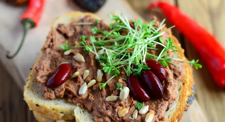 Pyszna pasta z czerwonej fasoli na chlebie, ozdobiona kiełkami i nasionami. Obok leżą papryczki chili.