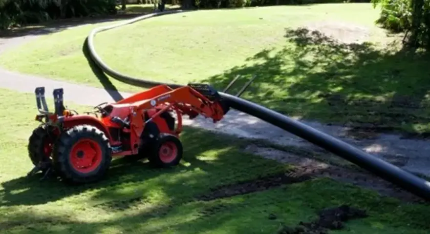 A tractor with a front loader is laying a long, flexible HDPE pipe across a grassy area.