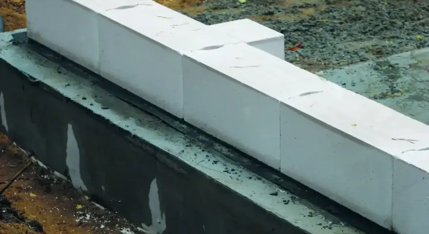 Białe bloczki betonowe układane na warstwie izolacji. Widać wilgotną zaprawę i ziemię wokół fundamentów.