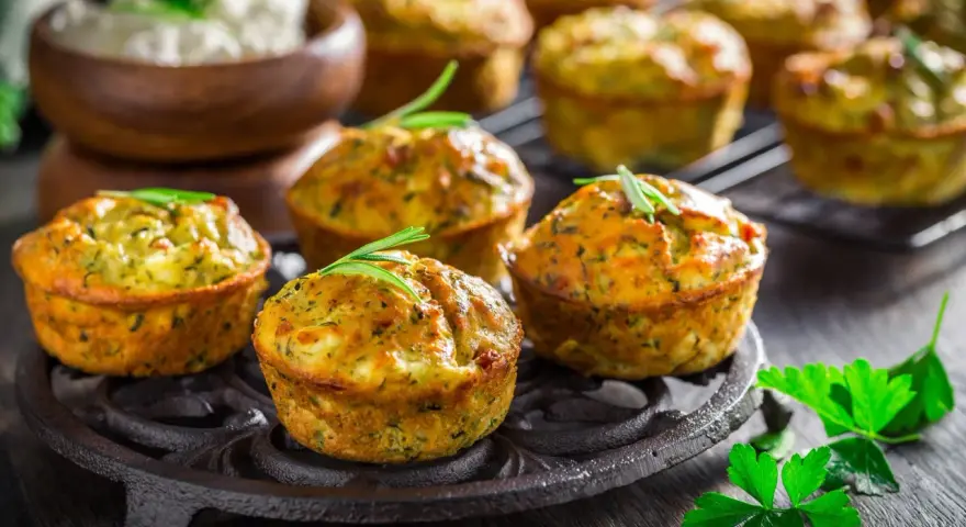 Deliziosi tortini di zucchine dorati, guarniti con rosmarino, su un piatto di ghisa. Sullo sfondo, una ciotola di formaggio fresco.