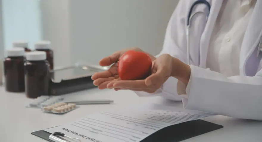 Lekarz trzyma w dłoniach czerwone serce, symbolizujące zdrowie. Na stole leżą leki i dokumentacja medyczna, sugerując rozmowę o tym, po jakim czasie spada cholesterol.