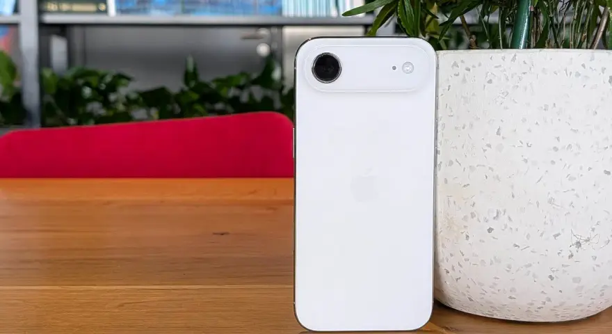 Biały iPhone z 4 aparatami, leżący na drewnianym blacie obok doniczki.