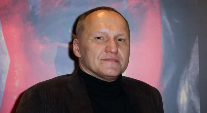 Portret Mariusza Grzegorzka w czarnym golfie i brązowej marynarce na tle abstrakcyjnego wzoru.