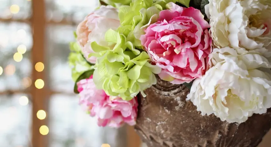 En bukett med rosa blomma, vita pioner och ljusgröna hortensior i en antik urn. Bakgrunden är mjukt suddig med ljus.