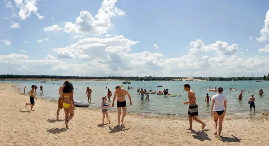 Menschen genießen einen sonnigen Tag am Badesee Frankfurt. Kinder planschen im Wasser, während Erwachsene sich am Sandstrand entspannen.