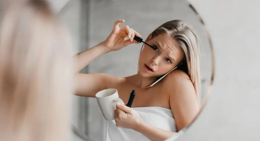 Une femme, enroulée dans une serviette, applique son mascara tout en tenant une tasse et en parlant au téléphone. Le mascara qui coule sur son visage témoigne de sa matinée mouvementée.