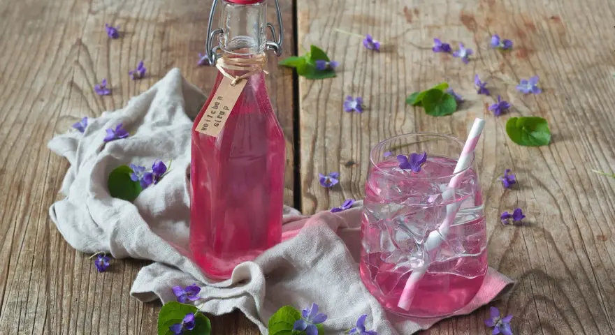 Flasche mit Veilchensirup und ein Glas mit Eis und Veilchenblüten auf Holzhintergrund.