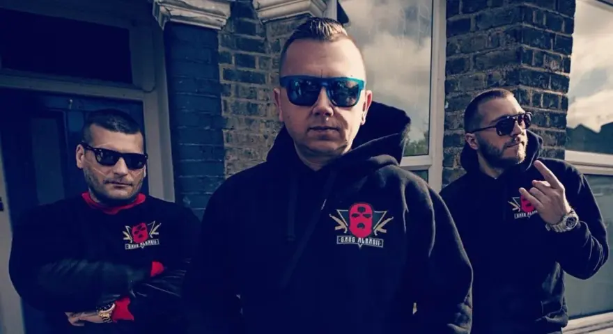 Trzech mężczyzn w bluzach z logo "Gang Albanii". Jeden z nich pokazuje gest ręką.