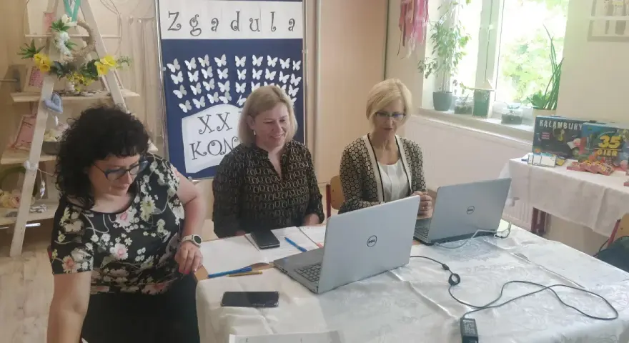 Trzy kobiety pracują przy stołach w sali, która wygląda jak szkoła specjalna. Na ścianie wisi tablica z motylami i napisem "Zgadula".