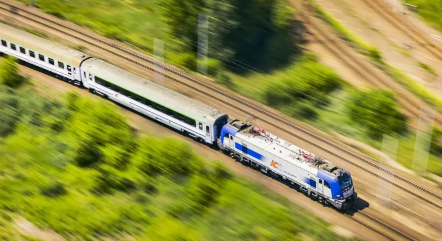 Pędzący pociąg PKP Intercity, model EU44 "Husarz", z białymi wagonami, mija zielone pola i drzewa.