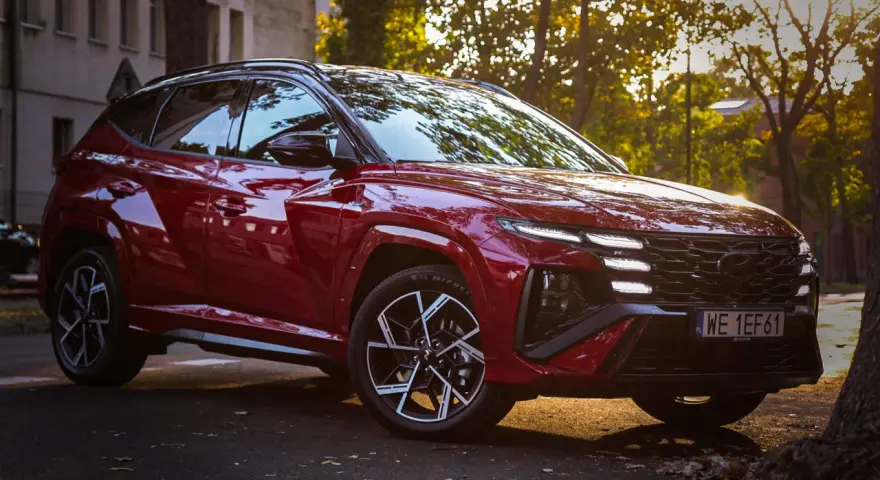Czerwony Hyundai Tucson Hybrid zaparkowany na tle jesiennych drzew i kamienicy.