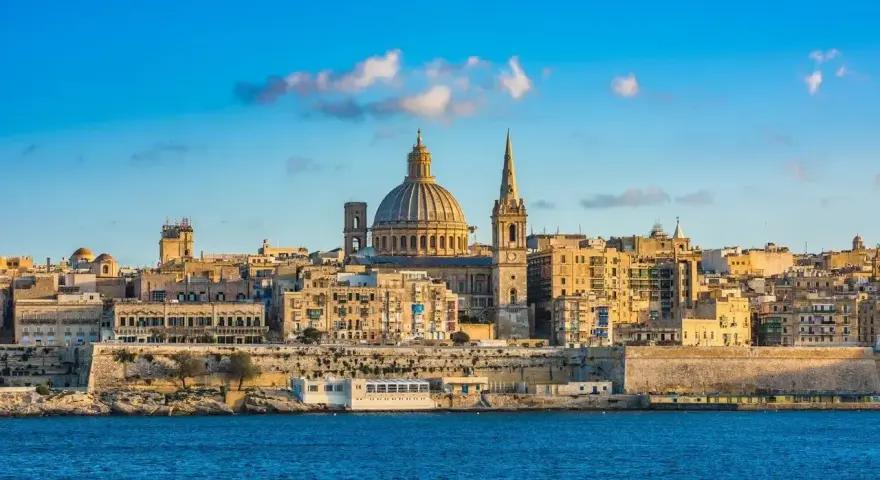 Malta, wyspa do kogo należy? Widok na Vallettę z kopułą i wieżą kościelną, otoczoną zabytkową architekturą, nad błękitnym morzem.
