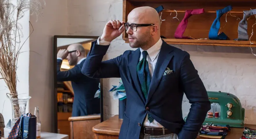 Elegancki mężczyzna w okularach dopasowuje krawat, tworząc idealny **zestaw koordynowany**. W tle widać kolorowe krawaty i poszetki.