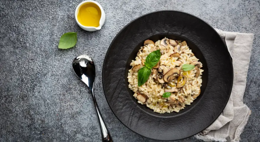 Kremowe risotto z grzybami na czarnym talerzu, obok łyżka i oliwa.