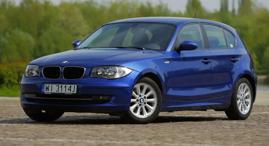Błękitne BMW E87 z polskimi tablicami WI 3114J, z nowymi żarówkami w reflektorach, zaparkowane na bruku.