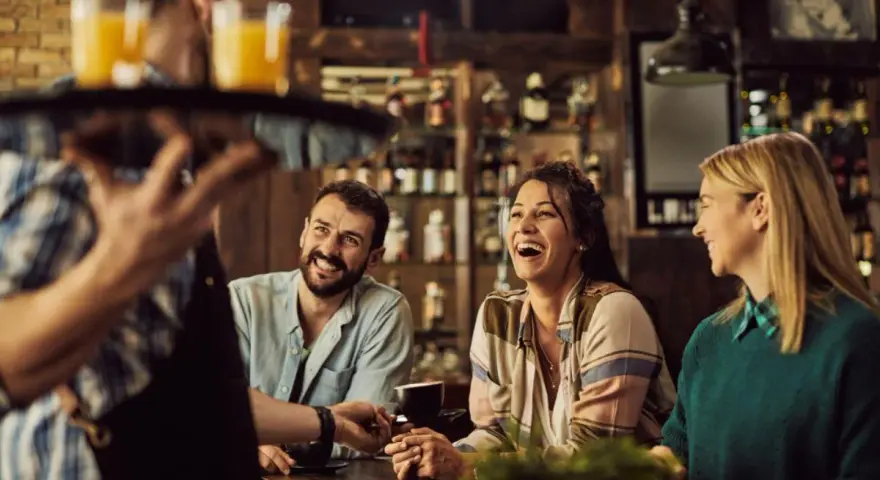 Un camarero sirve bebidas a un grupo de amigos riendo en un bar. La conversación animada sugiere que aún no es tarde para el ruido.