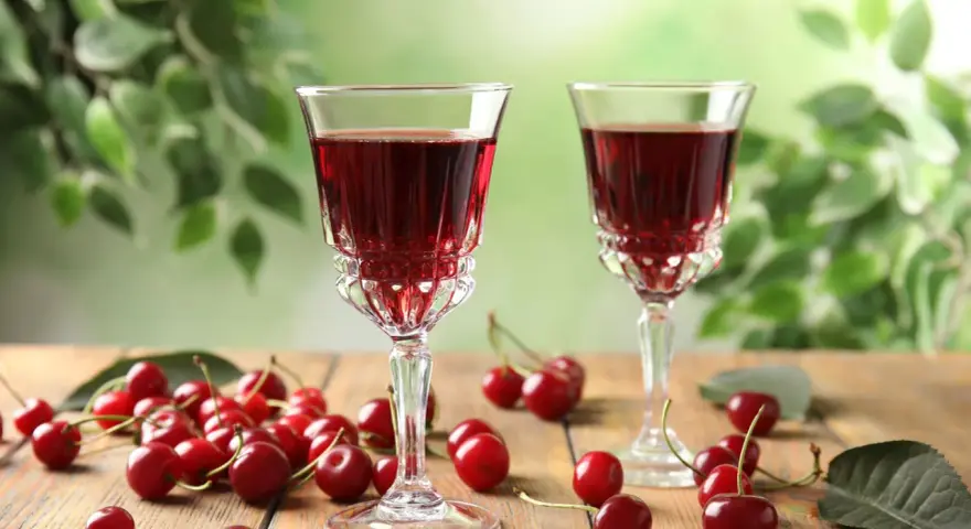 Dwa kieliszki z rubinową nalewką z wiśni na spirytusie, obok świeże wiśnie na drewnianym stole. Szybka nalewka z wiśni na spirytusie gotowa do degustacji.