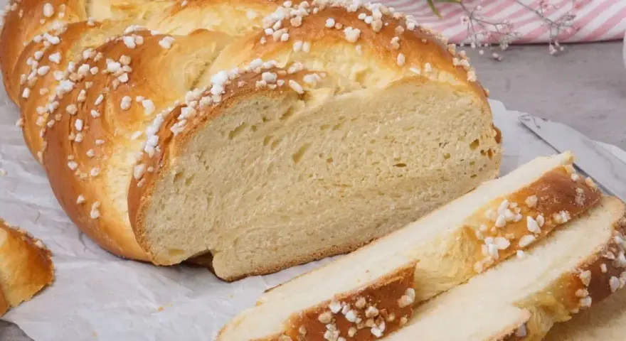 Ein goldbrauner Hefezopf, bestreut mit Zuckerkristallen, liegt auf Backpapier. Ein Stück ist angeschnitten, die weiche Krume ist sichtbar. Ein tolles Rezept für Hefezopf!