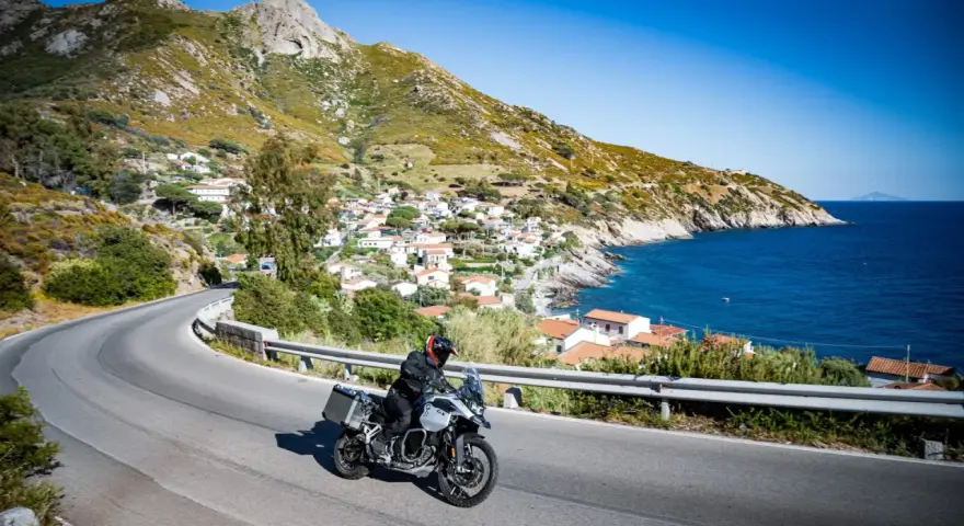 Un motociclista esplora le curve panoramiche dell'Elba, con il mare blu e un villaggio pittoresco sullo sfondo.