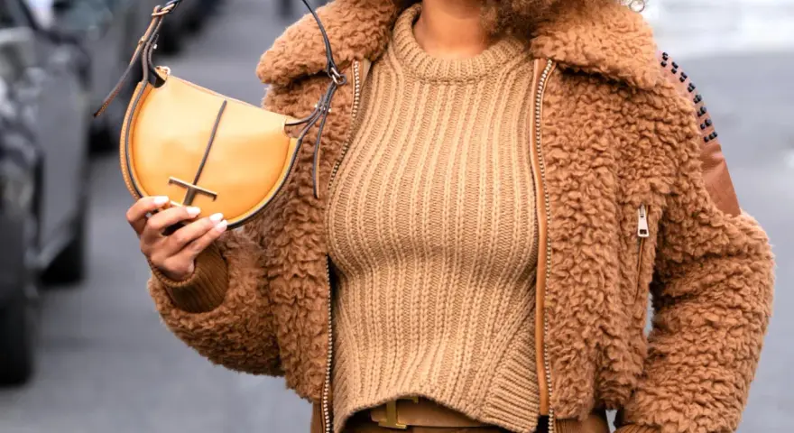 Une femme porte un pull beige côtelé, une veste en fausse fourrure marron et un sac à main jaune moutarde. Le jaune moutarde est une couleur qui va bien avec le beige.