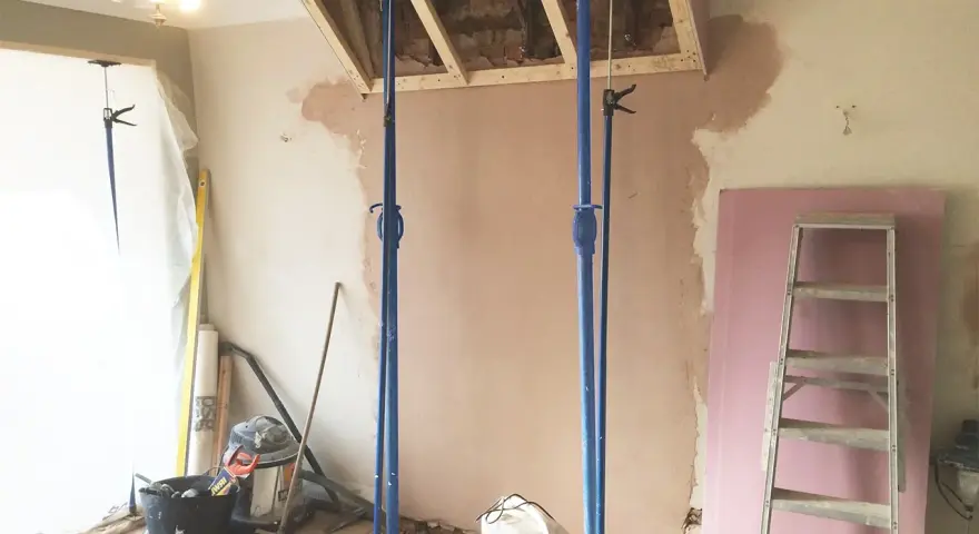 Travaux de rénovation : des étais soutiennent une structure en bois au-dessus d'une zone où l'on s'apprête à casser une cheminée. Matériaux de construction et outils au sol.