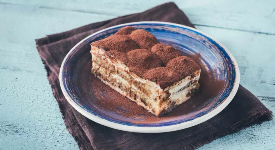Pyszny kawałek prawdziwego tiramisu, posypany kakao, na niebieskim talerzu.