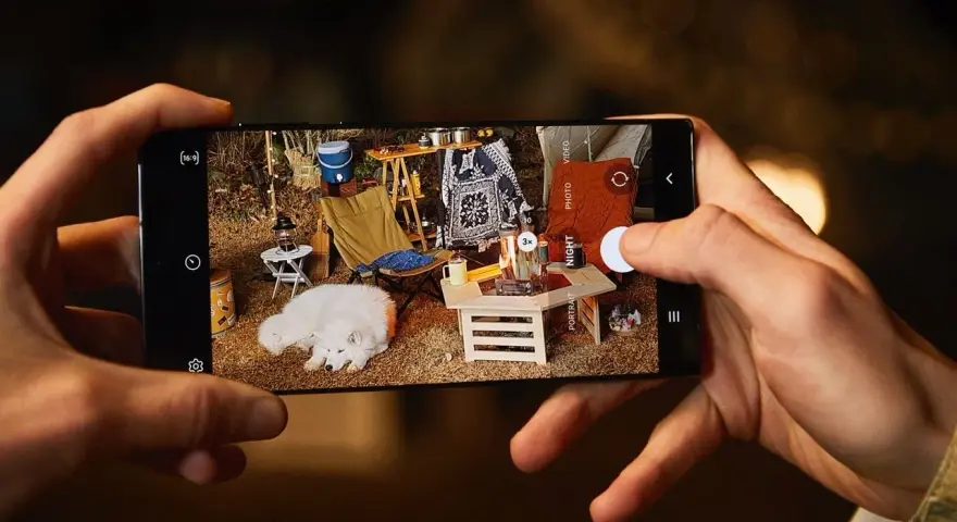 Une photo de téléphone capture un chien blanc endormi lors d'un camping. L'écran du téléphone montre une scène de camping avec une tente, une chaise pliante et un feu de camp.