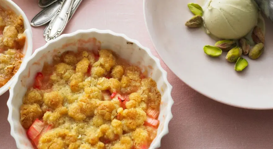 Rhabarber-Crumble in weißer Auflaufform, daneben eine Kugel Eis mit Pistazien. Ein köstliches Rhabarber Dessert.