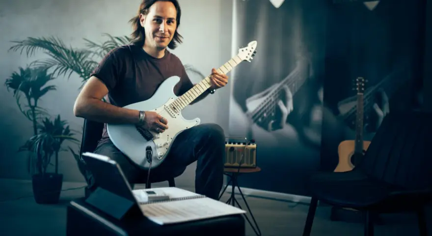Mann mit E-Gitarre, bereit, online Gitarre zu lernen. Noten und Tablet liegen bereit.