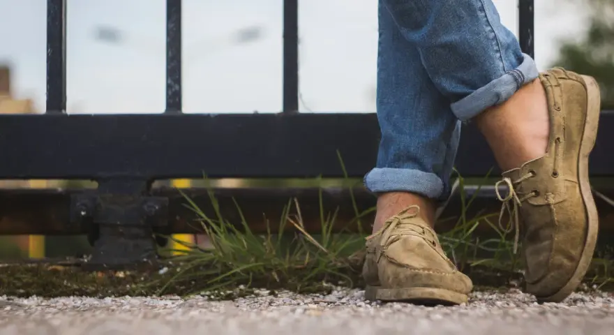 Gambe con jeans arrotolati e scarpe senza calzini, appoggiate su un muretto con erba.