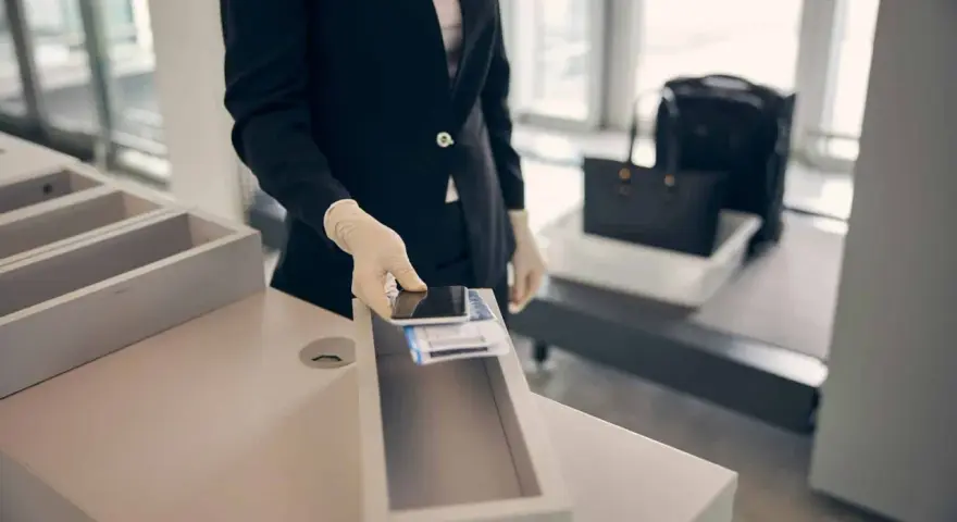 Pracownik lotniska w rękawiczkach kładzie telefon i karty pokładowe do pojemnika. W tle widać bagaż na taśmie. Zapalniczka w samolocie jest zabroniona.