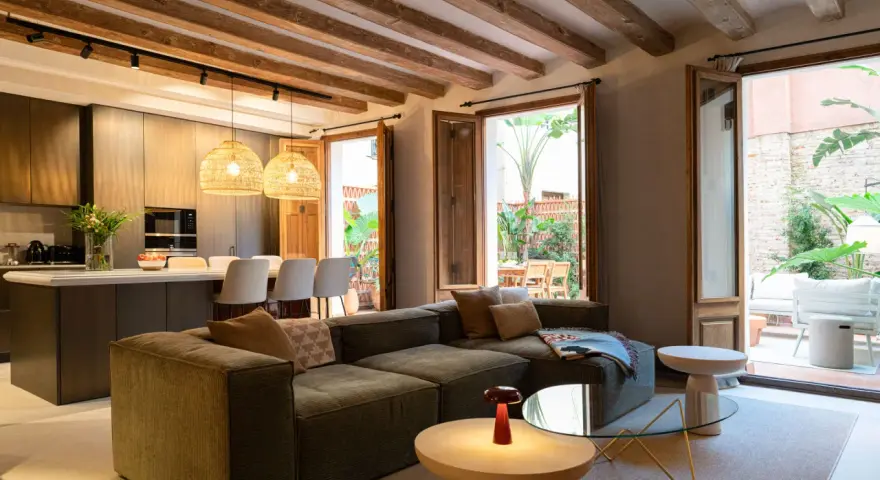Sofá modular verde con acabado fratasado, mesas auxiliares, lámpara roja y acceso a patio con muebles de exterior.