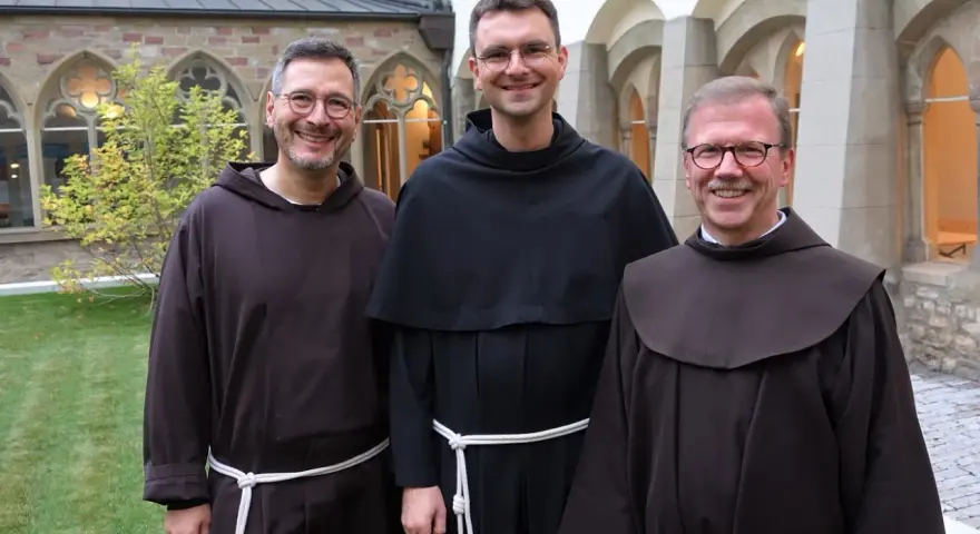 Drei Franziskaner Minoriten in braunen und schwarzen Kutten lächeln in die Kamera vor einer Klosterfassade.